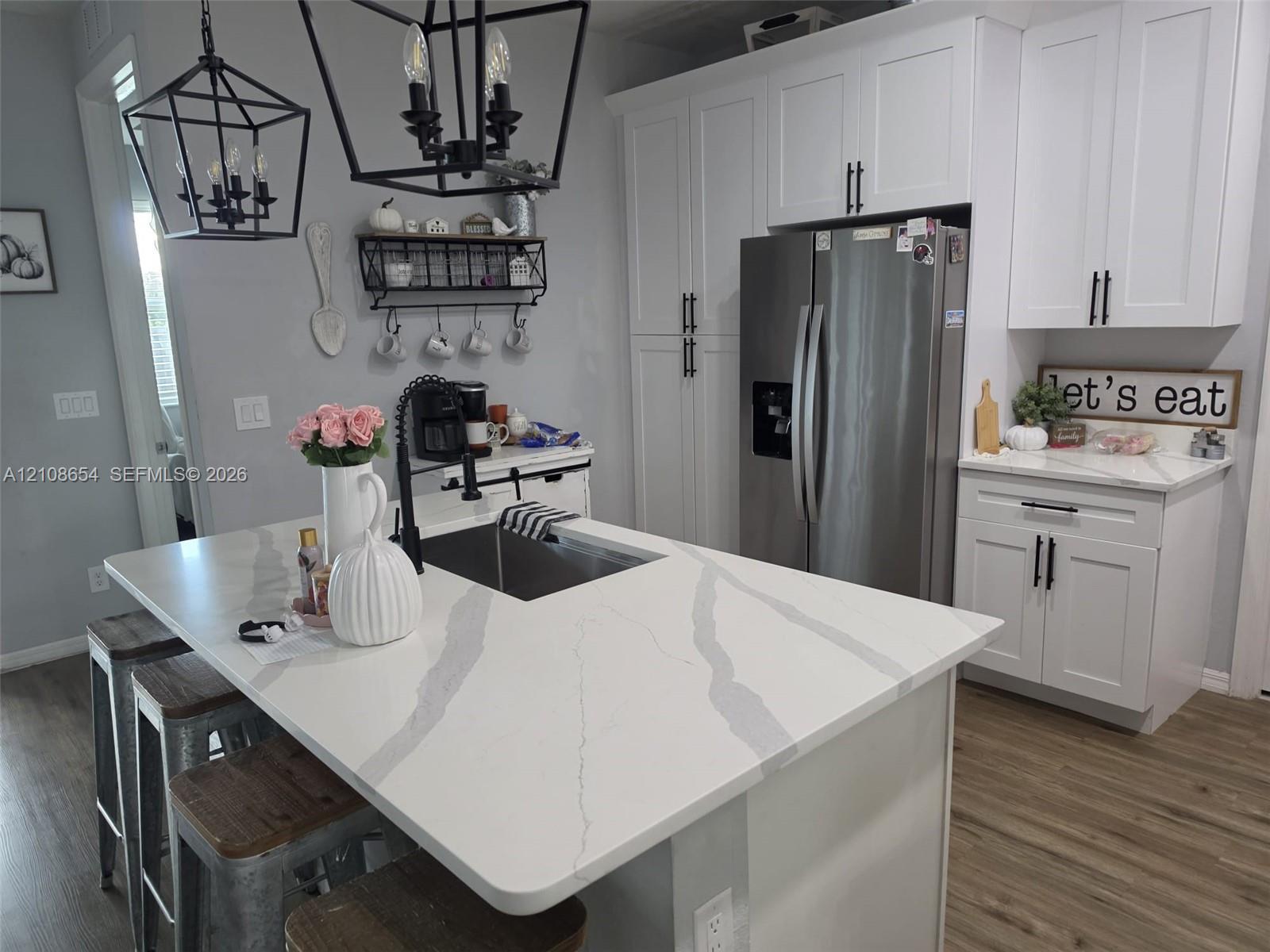 Kitchen featuring white shaker-style cabinetry, quartz countertops with veining, a central island with an integrated sink, geometric matte black pendant lighting, and wood-finish flooring