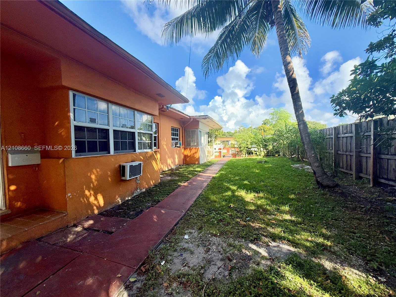 Side yard featuring a concrete walkway, exterior masonry wall, multi-pane windows, wood-style privacy fencing, and mature palm tree