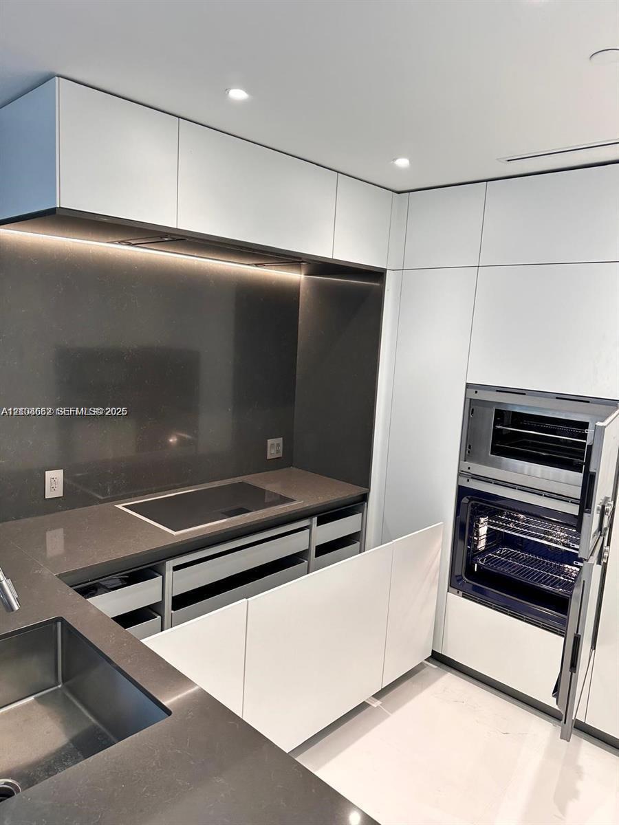 Modern kitchen featuring white flat-panel cabinetry, dark stone countertops, integrated induction cooktop, built-in dual oven, and undermount stainless steel sink