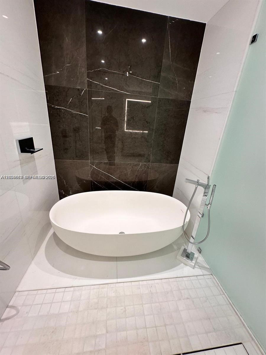 Freestanding oval soaking tub featuring a chrome floor-mounted faucet with a handheld sprayer, set against a dark large-format tile accent wall and white wall tile with a glass partition