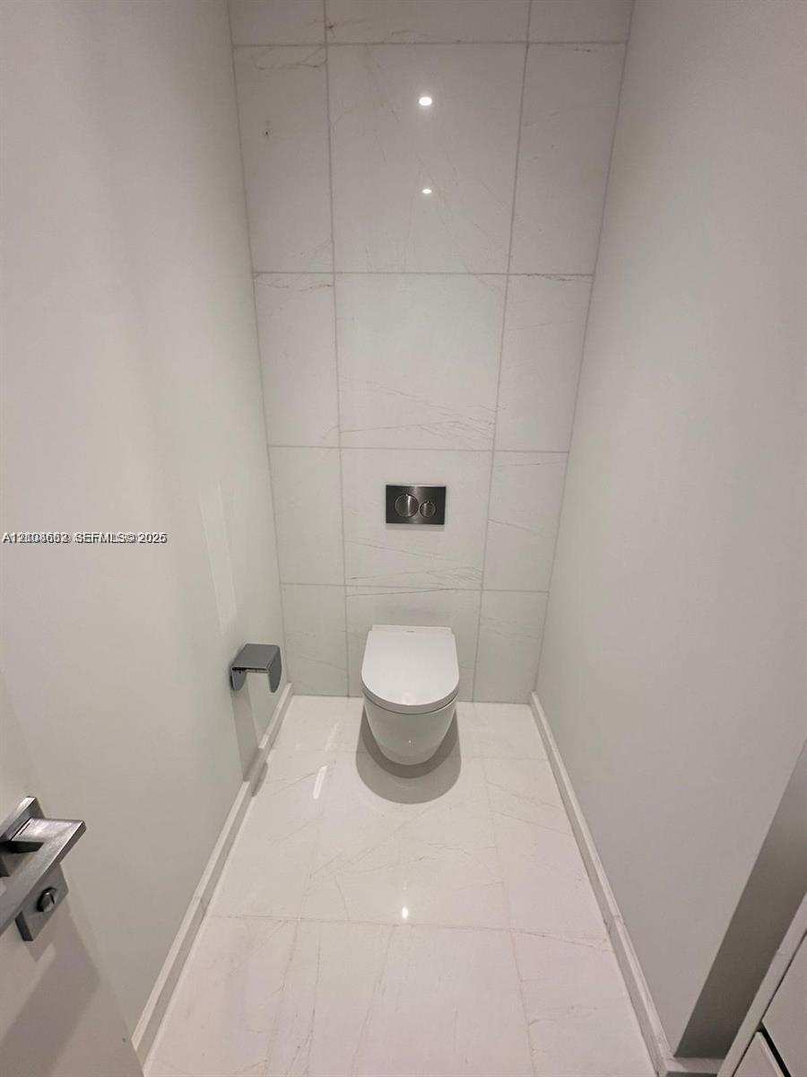 Wall-to-wall light-toned tile flooring, floor-to-ceiling tiled accent wall, wall-mounted toilet with a dual-flush actuator, and modern metal hardware