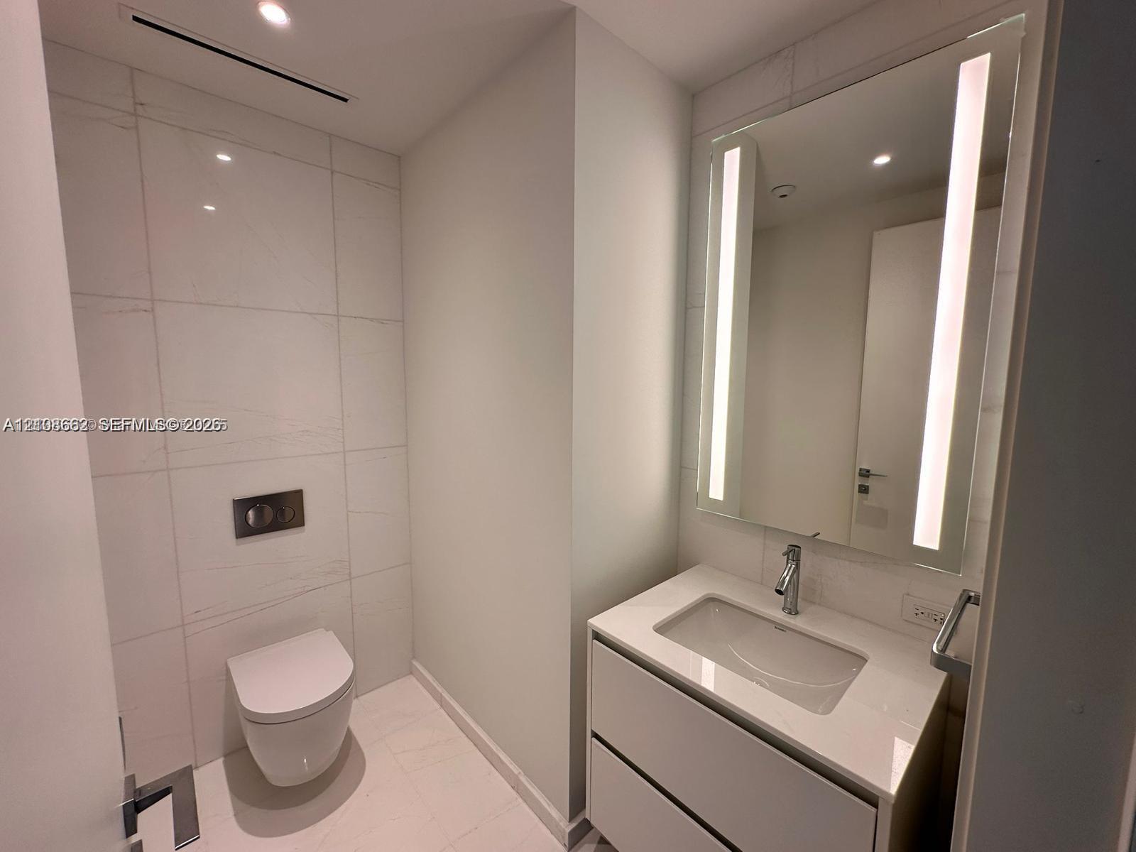 Wall-mounted toilet with a flush plate, vanity featuring a rectangular basin, integrated mirror with vertical LED lighting, and large-format porcelain tile wall cladding