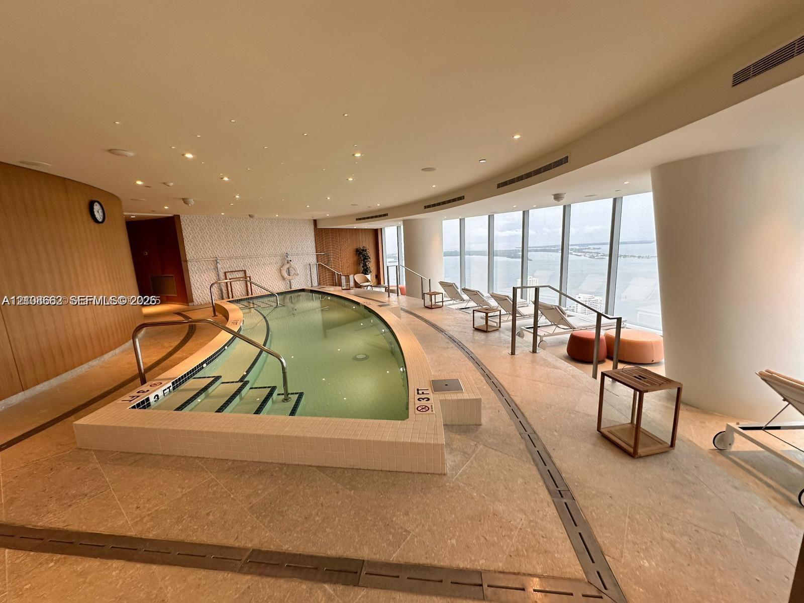 Indoor swimming pool featuring floor-to-ceiling windows, natural stone tile flooring, perimeter LED recessed lighting, wood-paneled walls, and built-in metal handrails