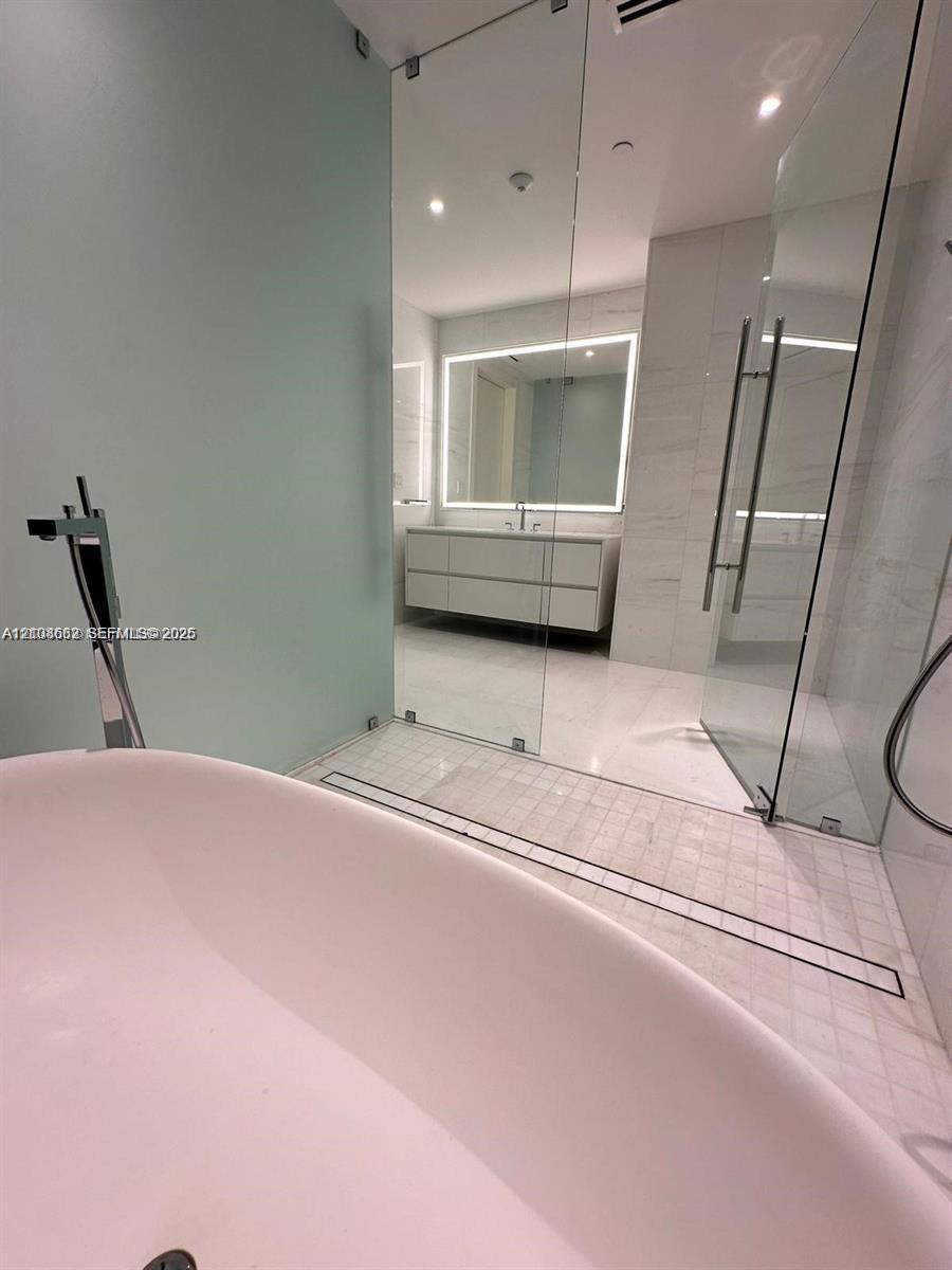 Freestanding soaking tub with a floor-mounted faucet, frameless glass-enclosed shower, mosaic tile shower flooring, wall-mounted double vanity with a backlit mirror, and large-format tile flooring