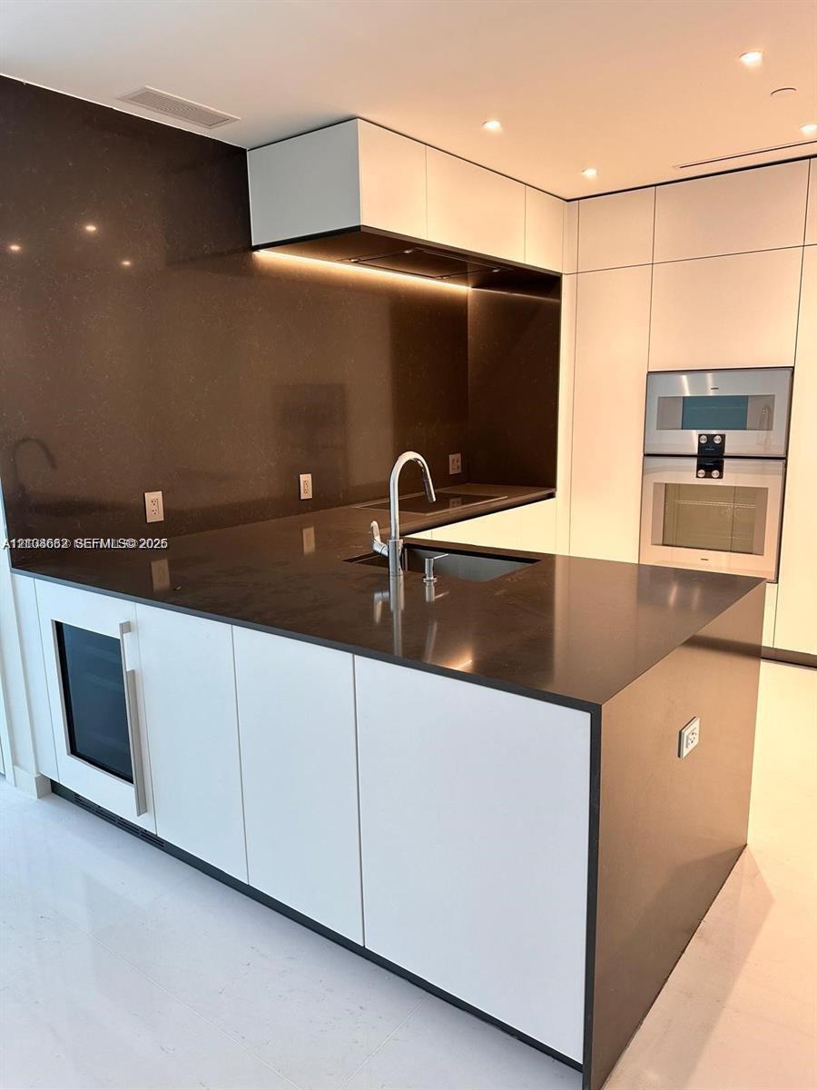 Modern kitchen featuring sleek white cabinetry, a dark stone waterfall-edge island, integrated stainless steel appliances, a flush-mount sink with a chrome faucet, and recessed lighting
