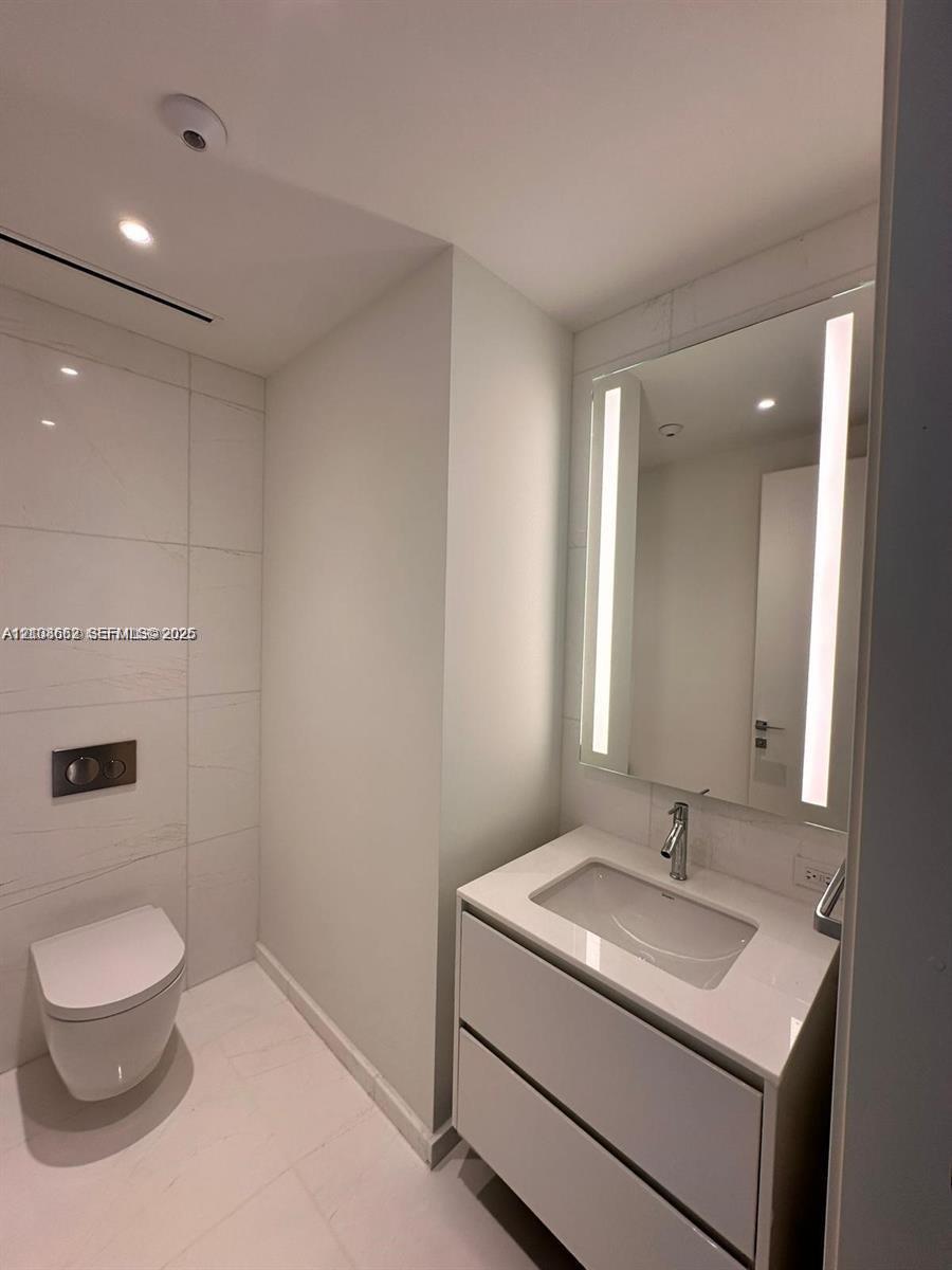 Modern bathroom featuring a wall-mounted toilet, floating vanity with a stone-finish countertop, integrated under-mount sink, chrome fixtures, and a vertical LED-backlit mirror