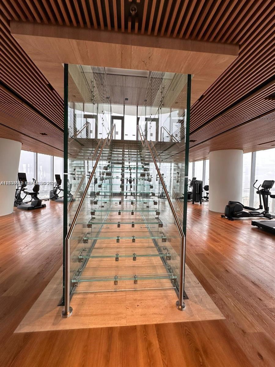 Staircase featuring glass treads and stainless steel railings, encased in a glass structural surround, complemented by a slatted wood ceiling, wood-finish flooring, and floor-to-ceiling windows
