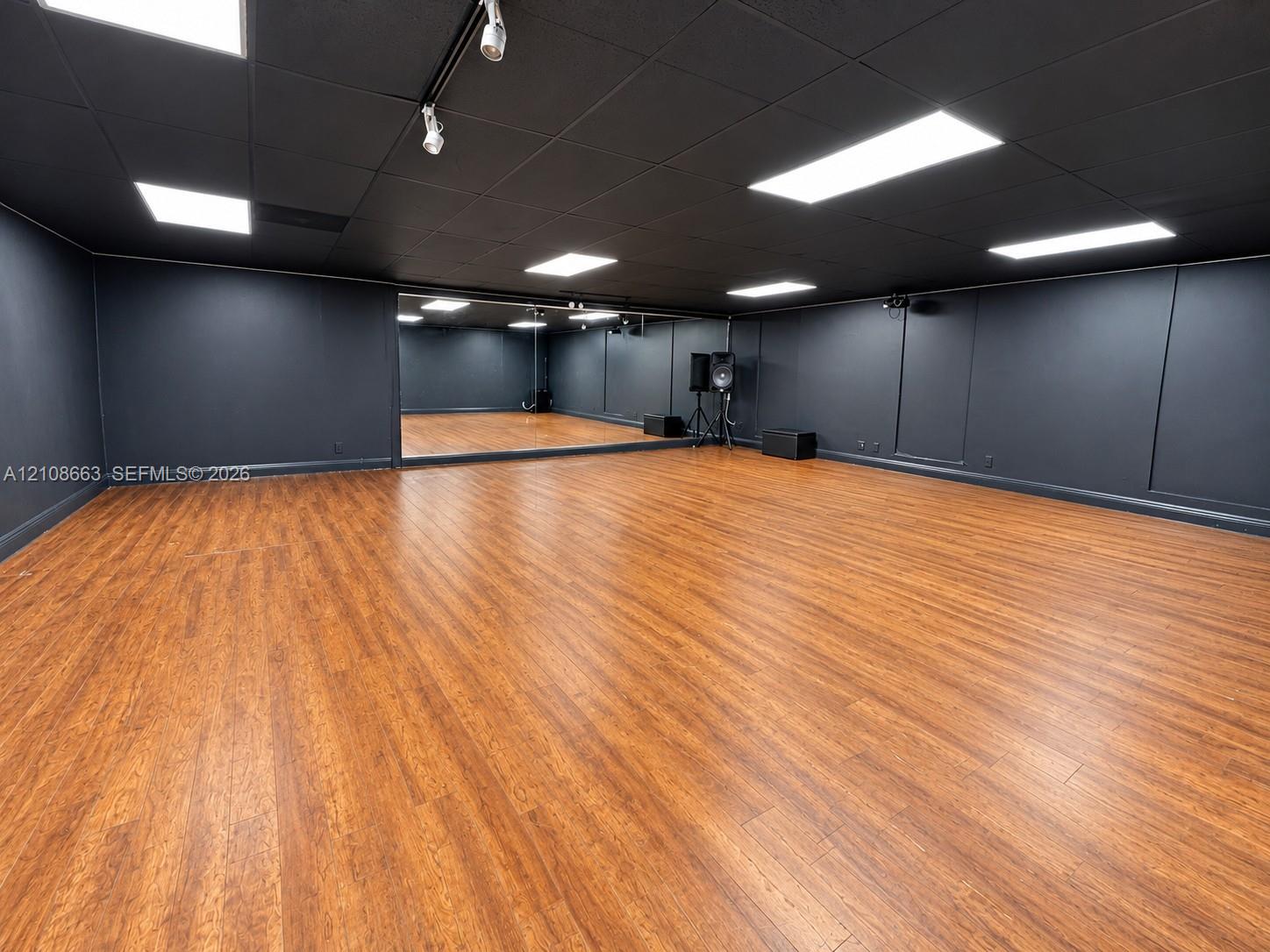 Expansive multi-purpose room featuring wood-finish flooring, full-wall mirrors, recessed lighting, and a dark-toned drop ceiling