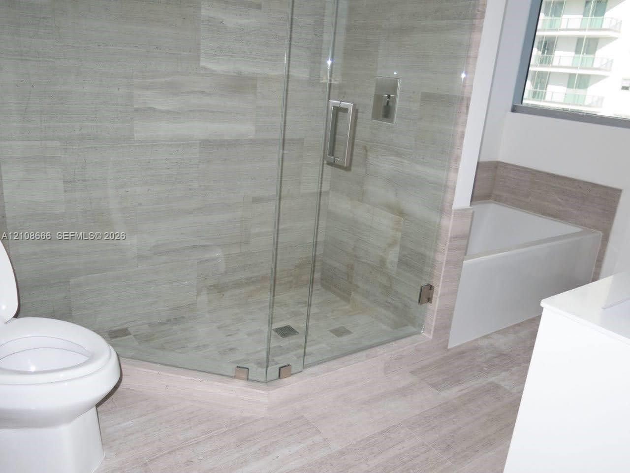 Walk-in glass-enclosed shower featuring large-format stone-finish tile surround and floor, deep soaking tub with tile surround, and neutral wood-finish flooring