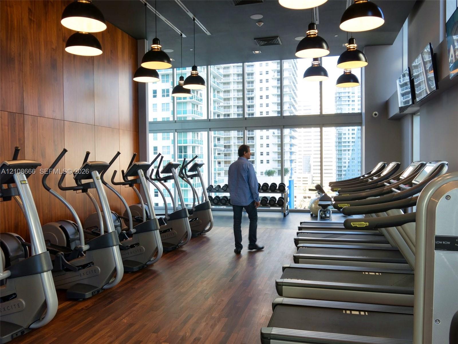 Fitness center featuring floor-to-ceiling windows, wood-finish flooring, wood-paneled accent wall, and industrial-style pendant lighting
