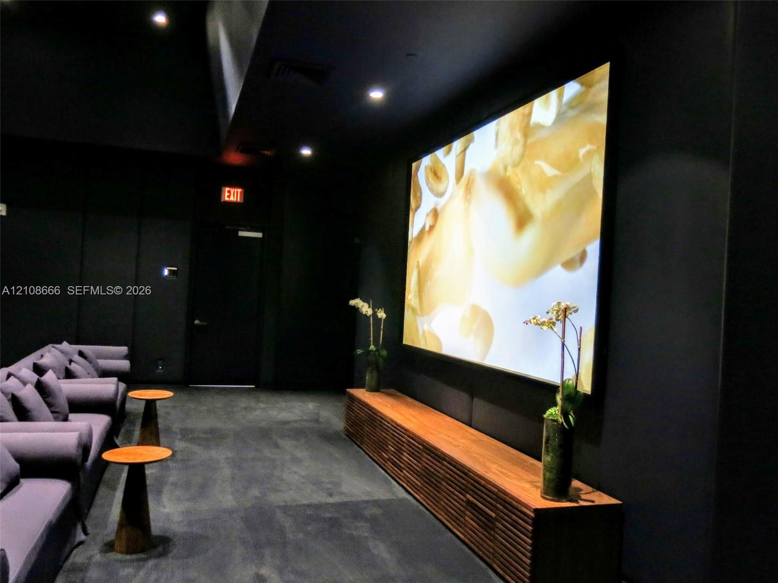 Media room featuring dark wall finishes, a large-format mounted screen, and a custom slatted wood-finish media console with recessed ceiling lighting