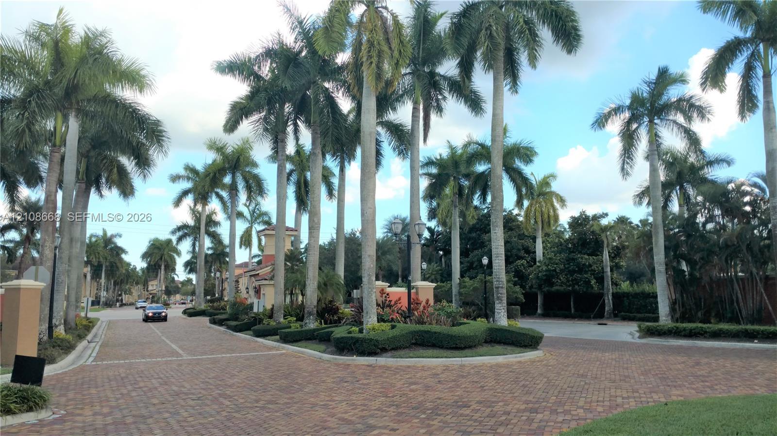 Paver driveway and road featuring mature palm trees, manicured landscape hedging, and decorative street lighting