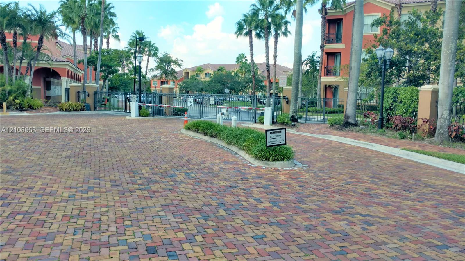 Gated community entry featuring paver-stone driveway, metal security gate, and mature palm trees