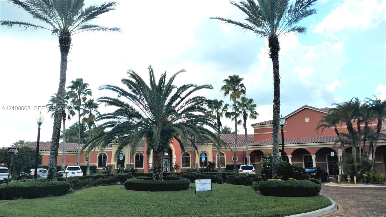 Stucco exterior featuring arched breezeways, a terra-cotta tile roof, mature palm trees, manicured landscaping, and a brick-paver circular driveway