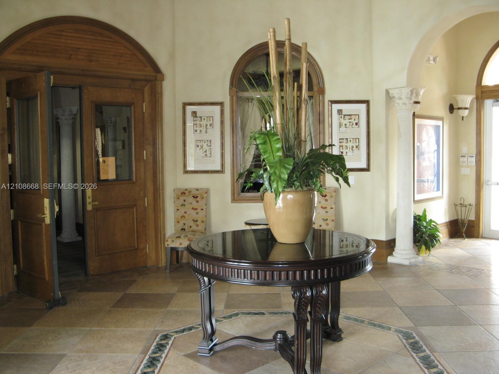 Foyer featuring arched wood double doors, decorative column, tile flooring with intricate border inlay, arched window, and wall-mounted sconce lighting