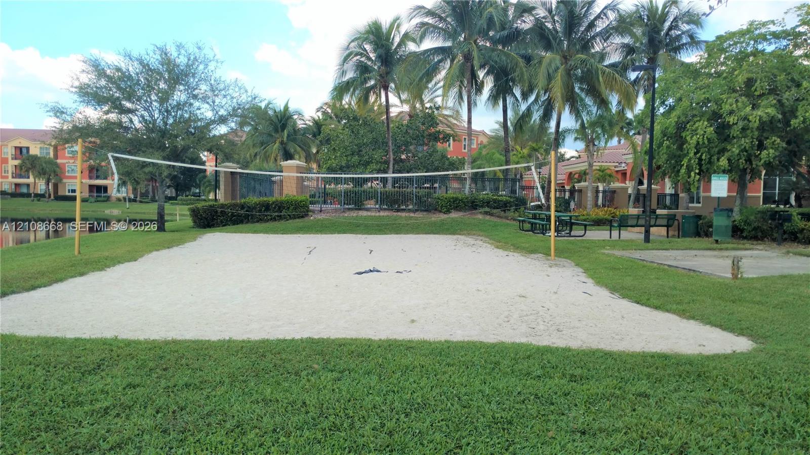 Sand volleyball court featuring a permanent net, manicured lawn, perimeter landscaping, and mature palm trees