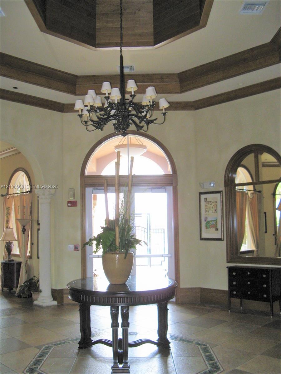 Foyer featuring a coffered octagonal ceiling with wood-finish trim, a prominent iron chandelier, arched doorways, tiled flooring with an inlaid decorative border, and a decorative column