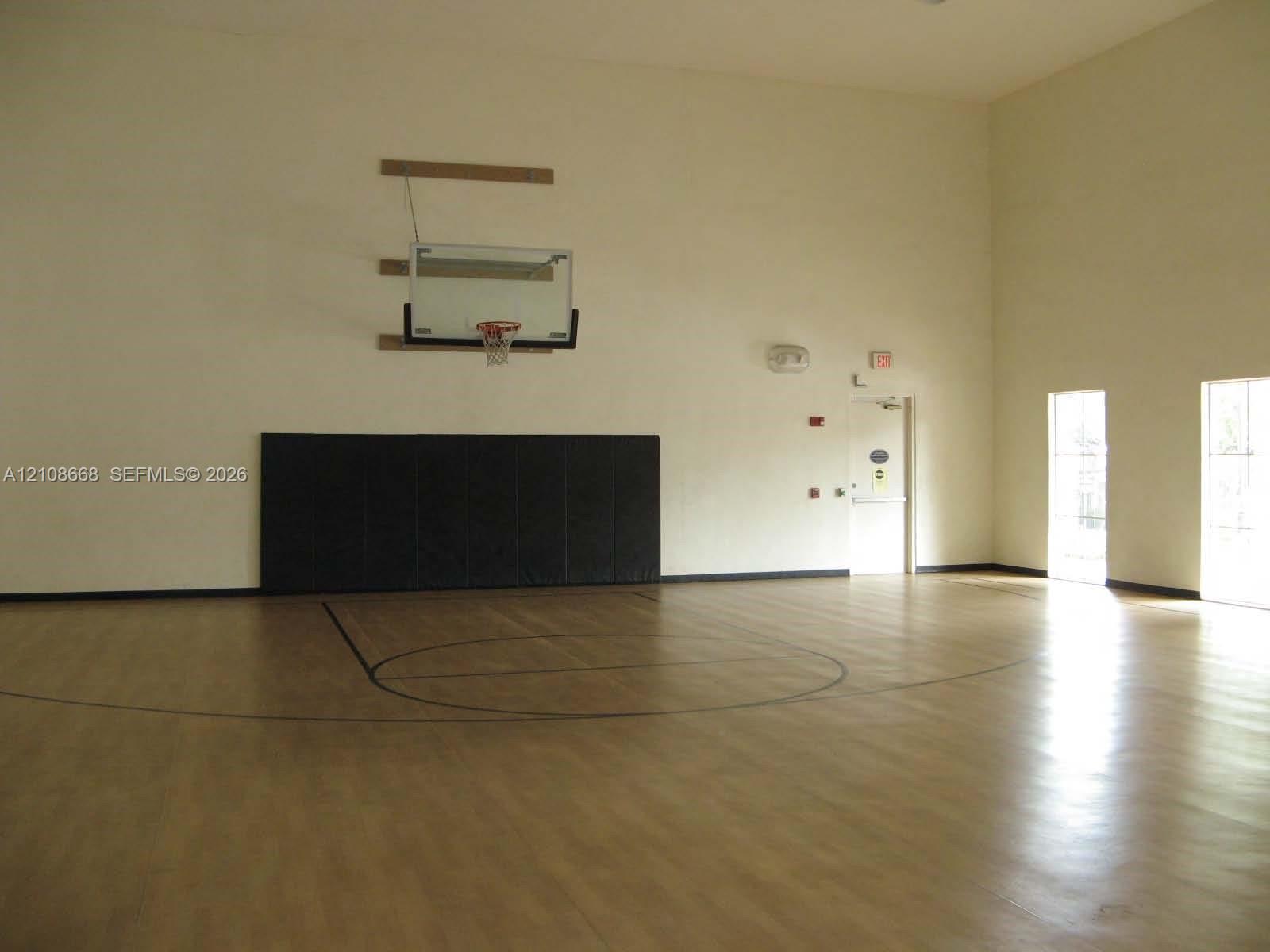 Indoor sports court featuring high ceilings, wood-finish flooring, a wall-mounted basketball hoop, and protective wall padding