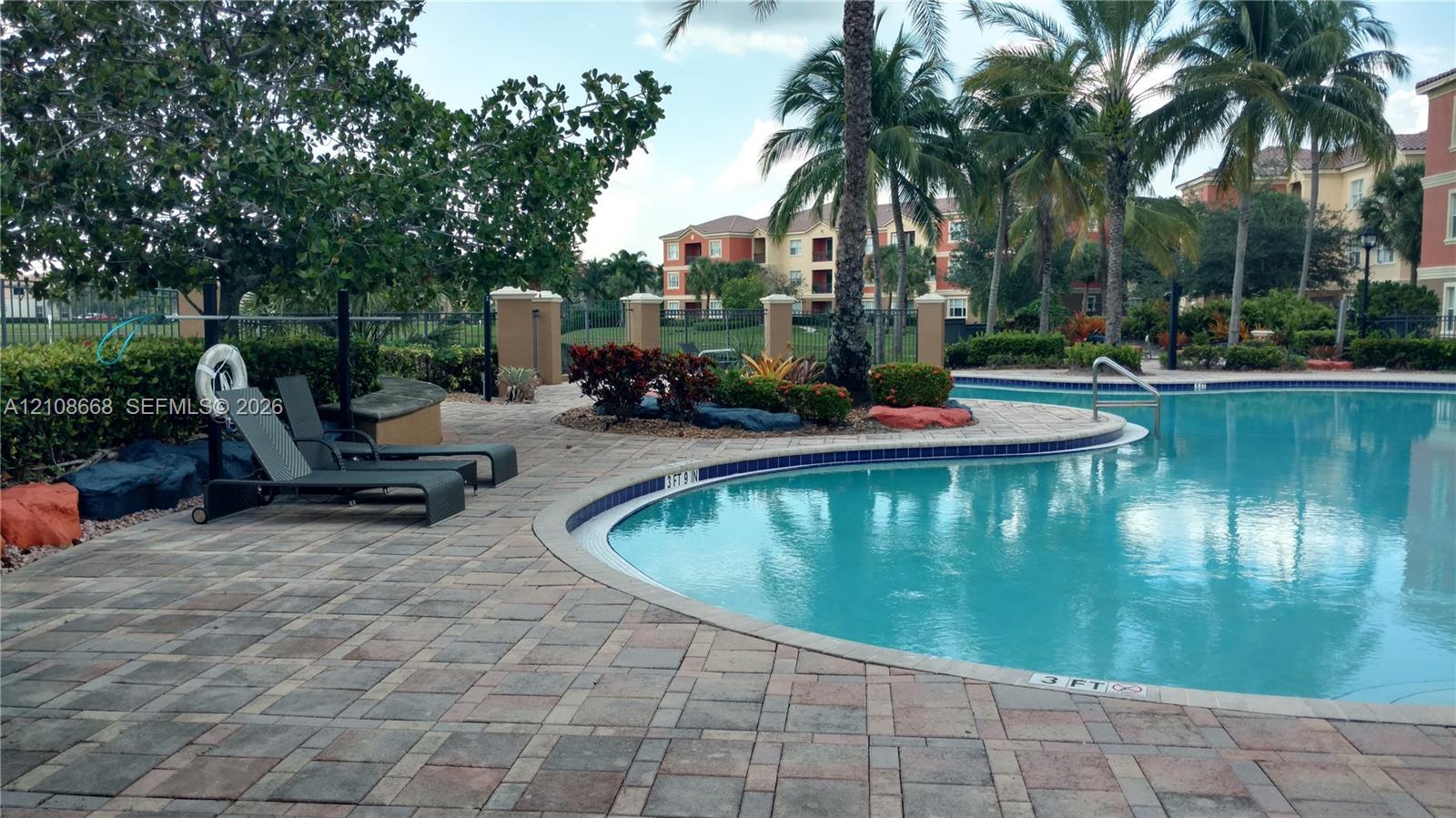 Curved swimming pool featuring a paver deck, mature palm trees, integrated landscaping, and decorative boulder accents