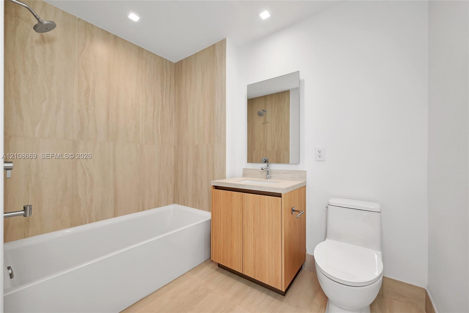 Bathroom featuring a wood-finish vanity with a stone countertop, a white soaking tub with floor-to-ceiling textured tile surround, a wall-mounted mirror, and recessed lighting