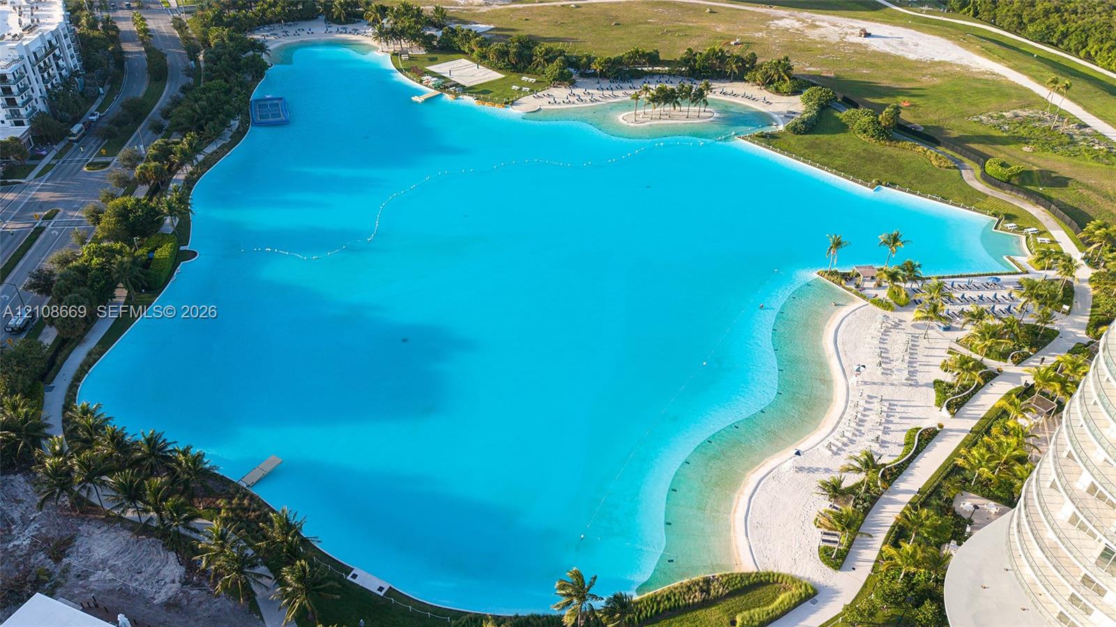 Expansive turquoise man-made lagoon featuring sandy beach shorelines, palm tree landscaping, paved walkways, and designated swimming areas