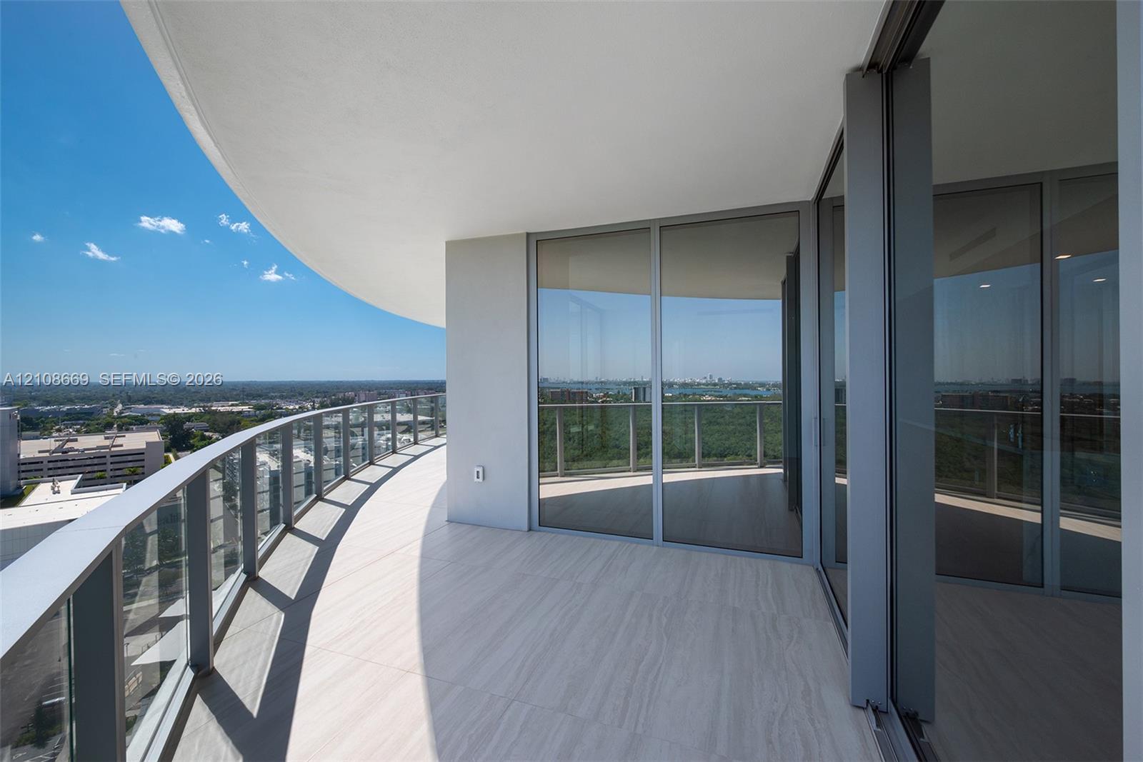 Curved wrap-around balcony featuring large-format tile flooring, glass-panel railing, and floor-to-ceiling sliding glass doors