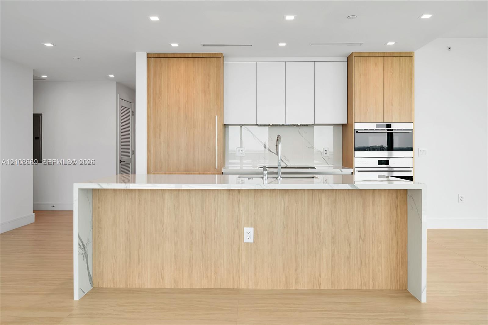 Kitchen featuring a waterfall-edge island, wood-finish cabinetry, white upper cabinets, stone-surfaced countertops and backsplash, and built-in stainless steel appliances
