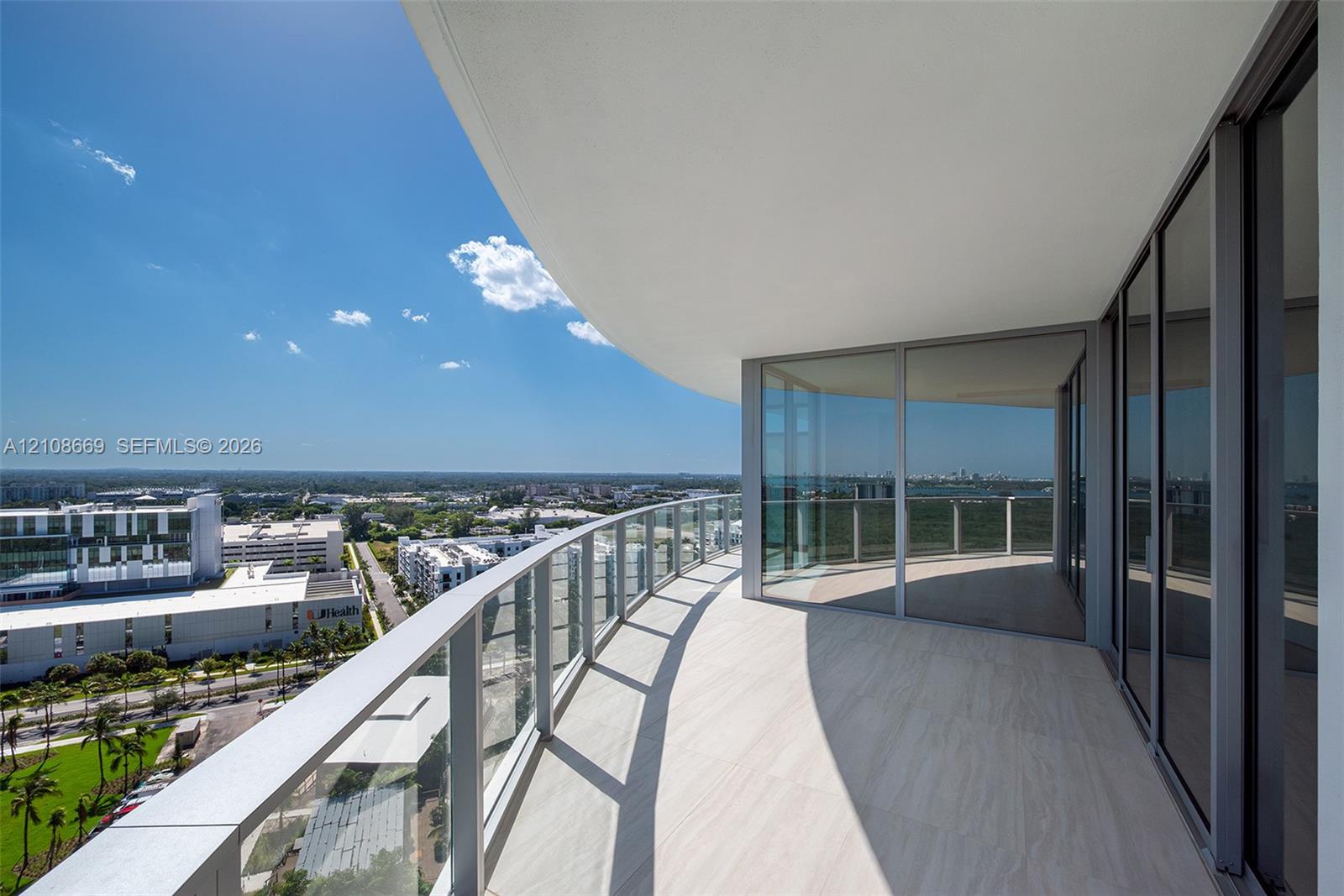 Expansive wraparound balcony featuring sleek glass railings, overhead recessed lighting, and large-format tile flooring, complemented by floor-to-ceiling glass sliding doors