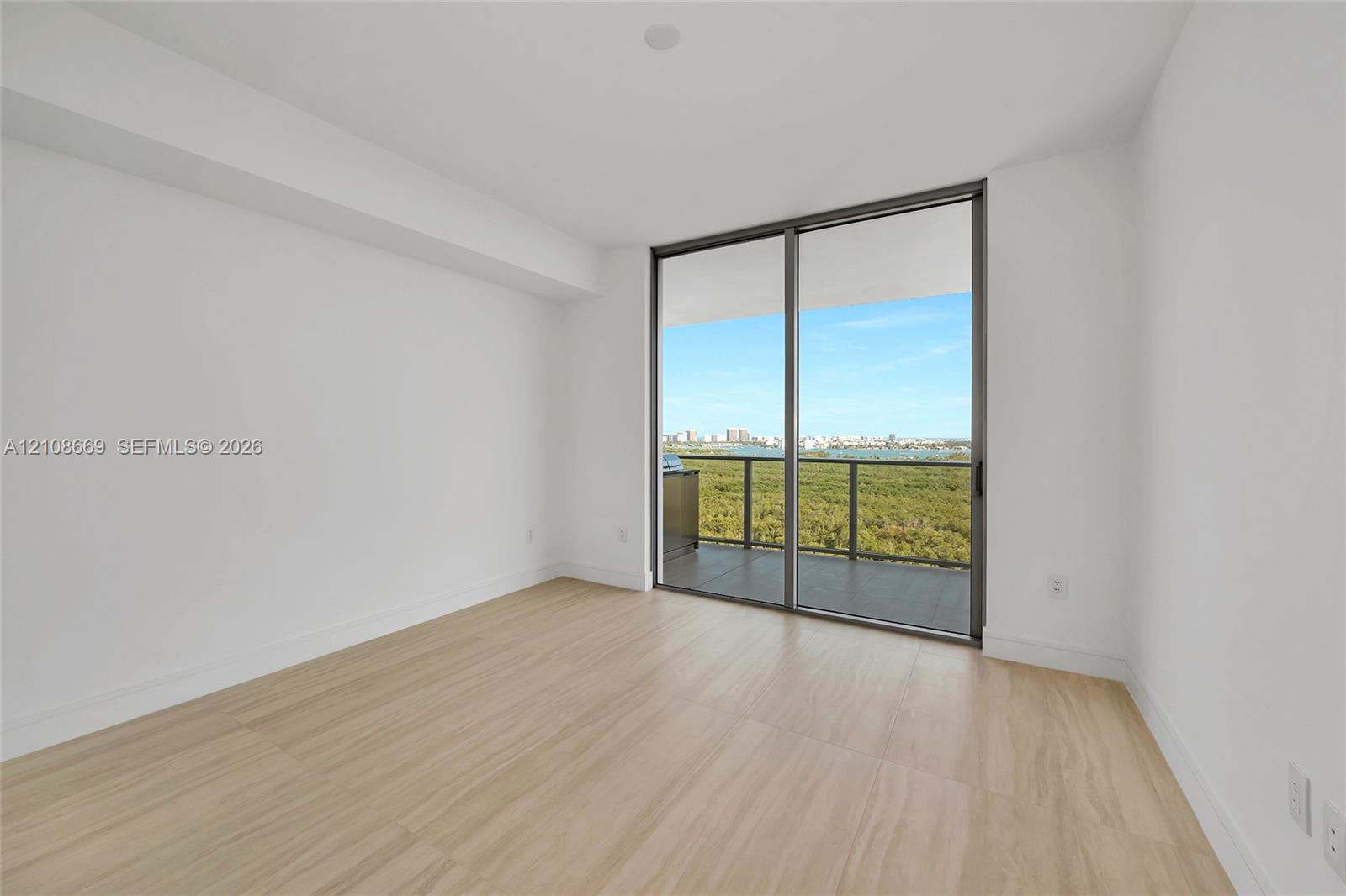 Neutral-toned interior featuring wood-finish flooring, full-height floor-to-ceiling glass sliding doors, and a private balcony with water and coastal scenery
