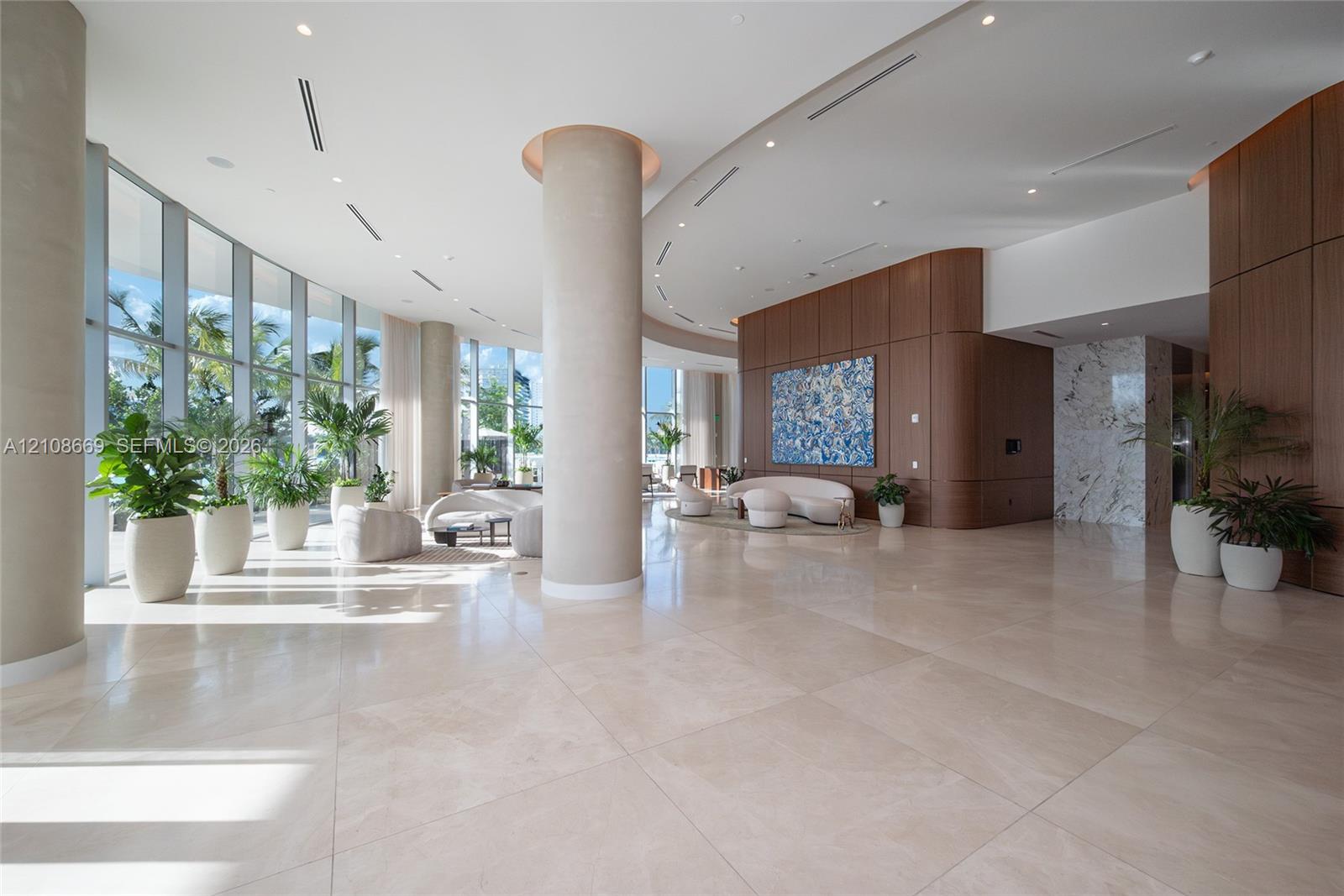 Expansive lobby featuring floor-to-ceiling glass walls, large-format tile flooring, wood-paneled walls, and structural columns with recessed lighting