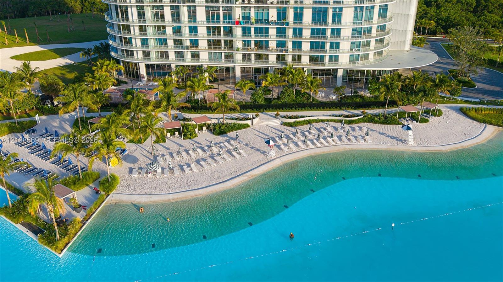 High-rise residential building with curved glass facade, expansive sand beach, lagoon-style pool, shaded cabanas, and mature palm landscaping