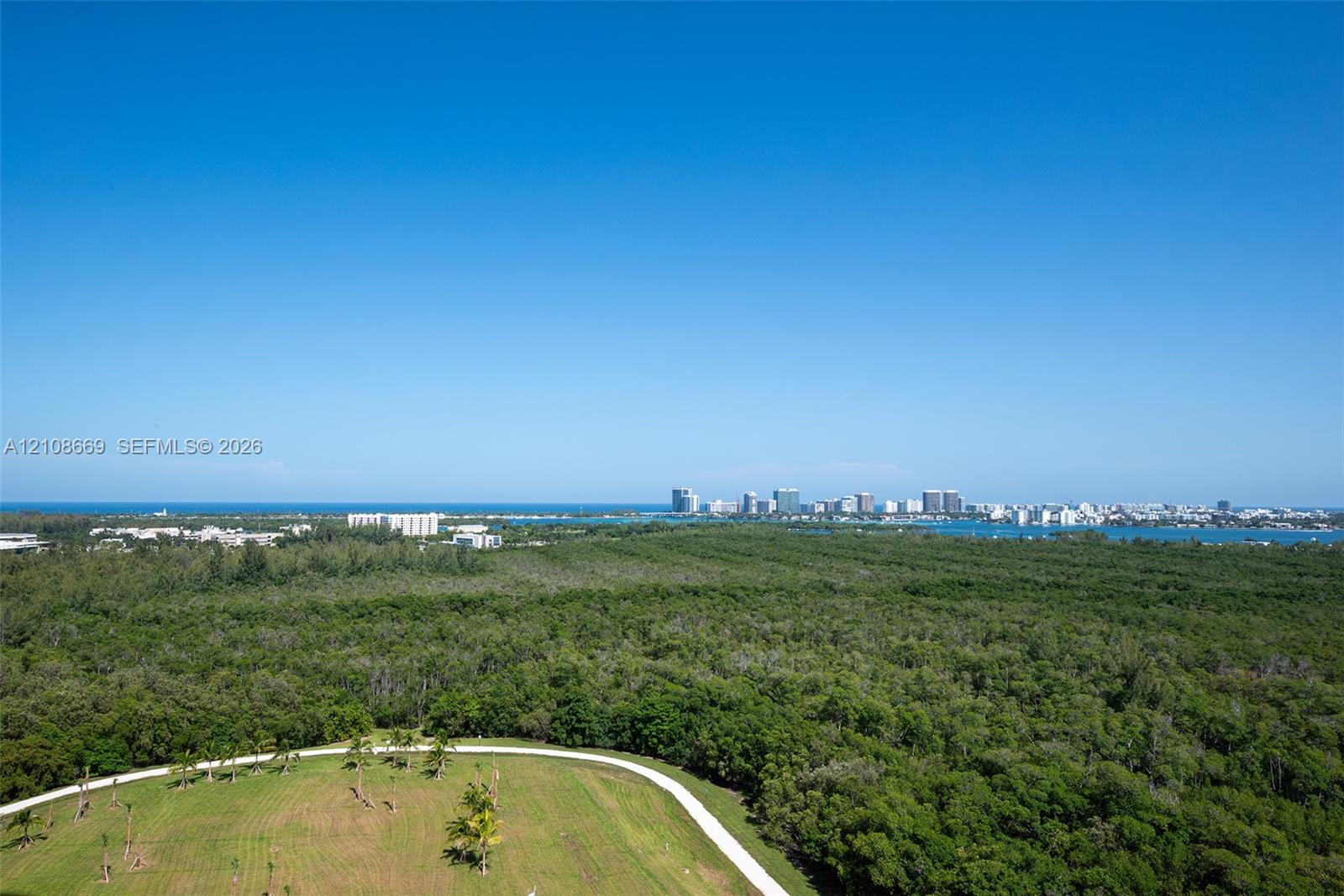 Aerial perspective showcasing extensive natural greenery, open lawn space, paved pathways, coastal water frontage, and a distant cityscape