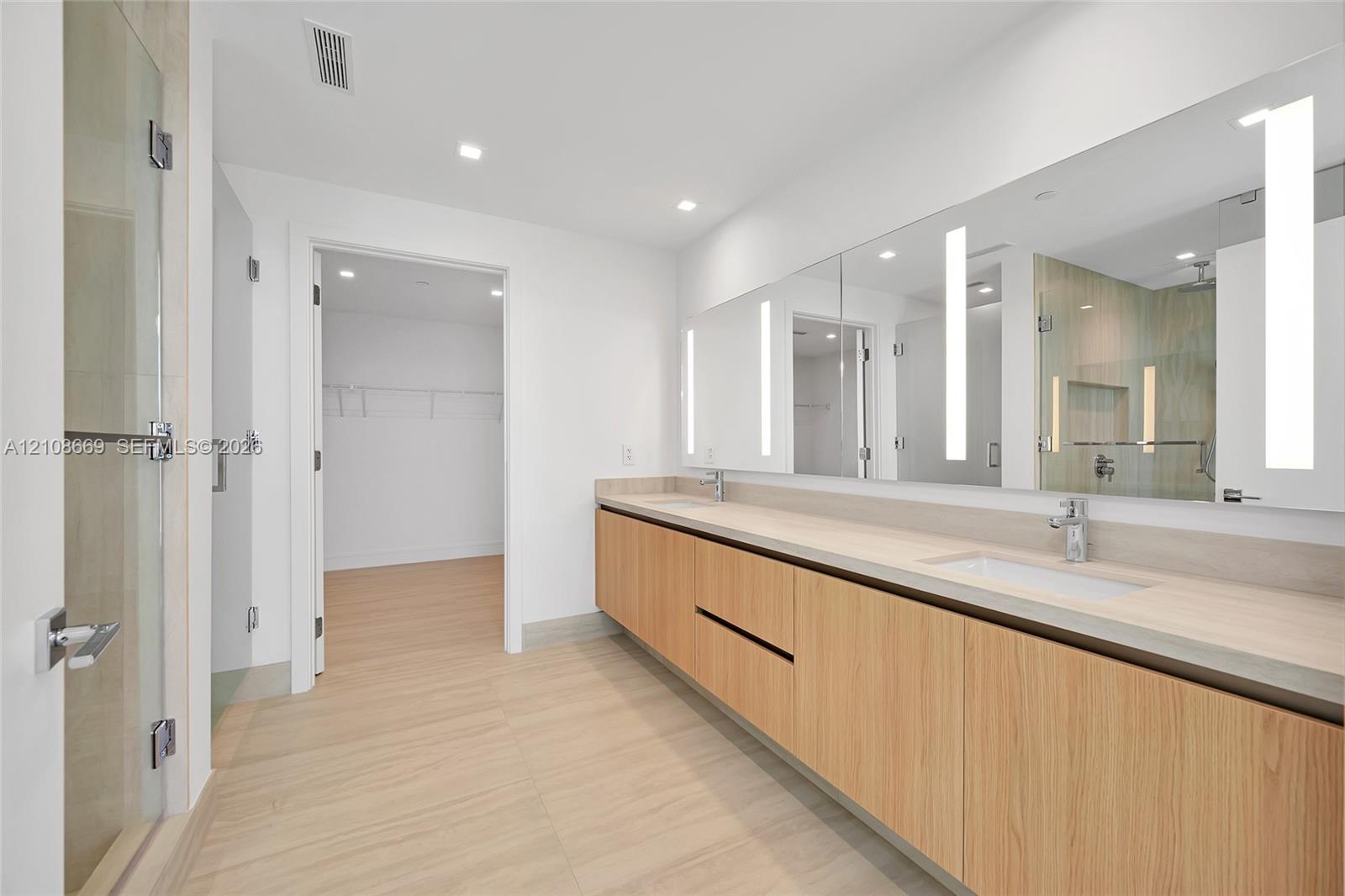 Dual-sink vanity with wood-finish cabinetry, countertop with integrated rectangular basins, large wall-mounted mirrors with integrated vertical lighting, and a glass-enclosed shower