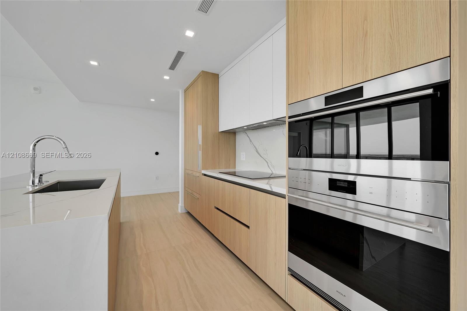 Kitchen featuring light wood-finish cabinetry, quartz countertops with a veined pattern, integrated stainless steel double wall ovens, a sleek electric cooktop, and a kitchen island with an undermount sink and chrome faucet