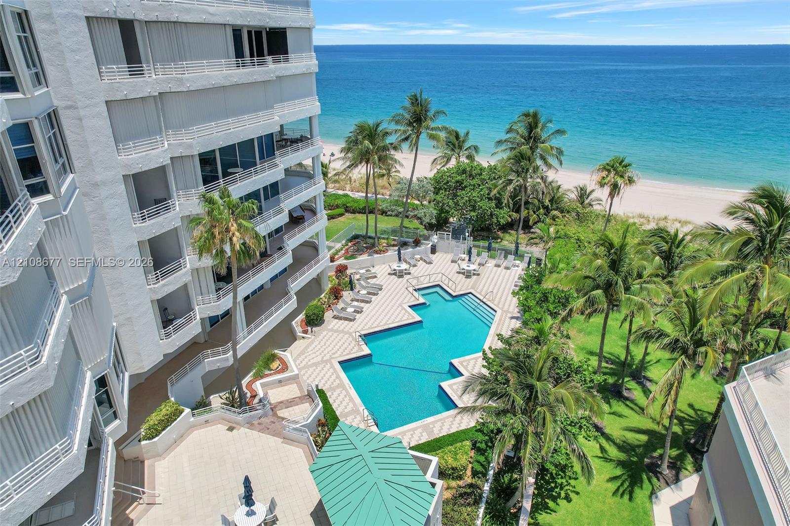 Multi-story residential building with private balconies overlooking an outdoor swimming pool, paved pool deck, manicured grounds with palm trees, and direct oceanfront access