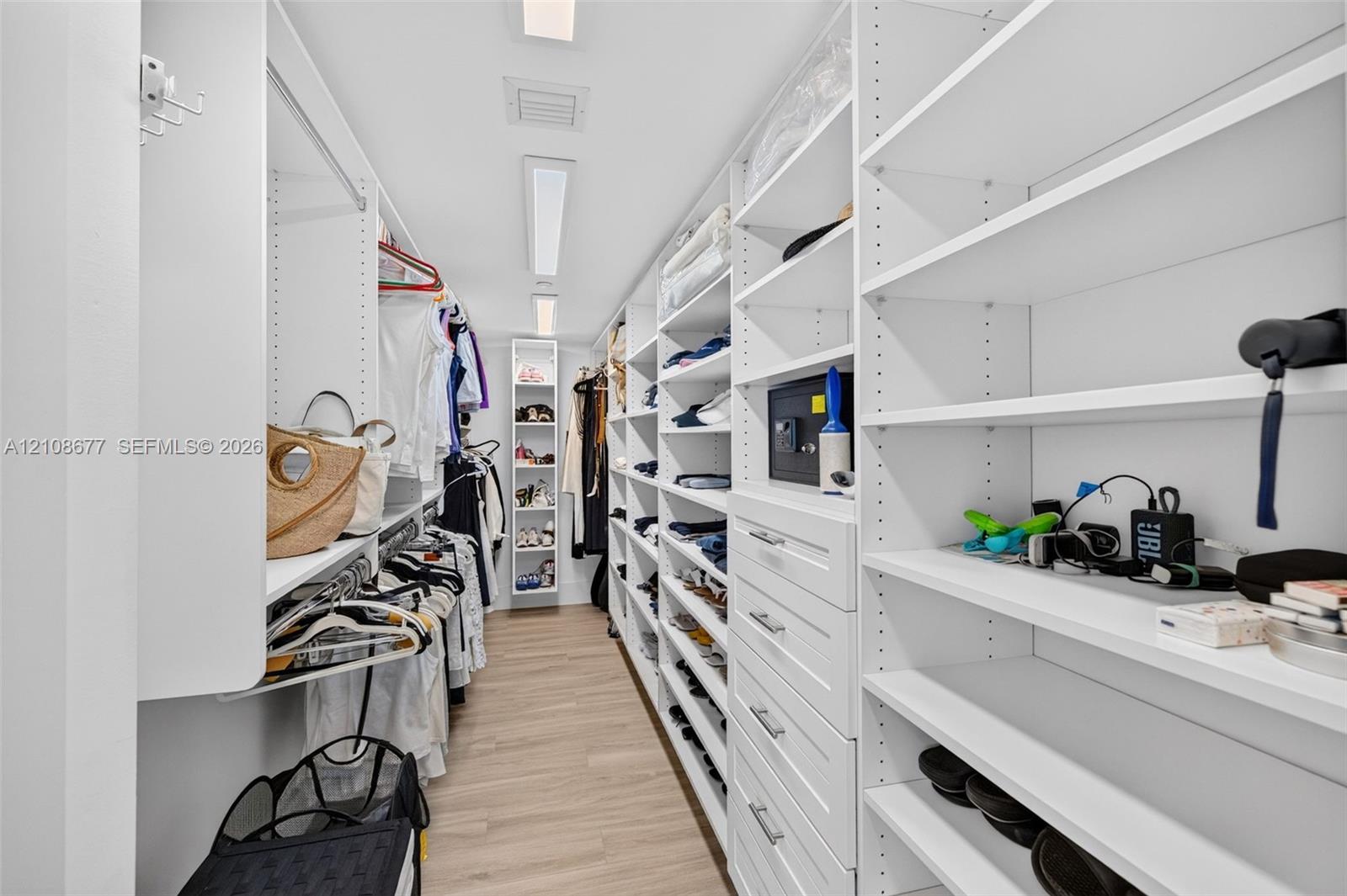 View toward long side of the custom walk-in closet featuring floor-to-ceiling white shelving, integrated cabinetry with drawers, multiple hanging rods, and wood look plank porcelain flooring