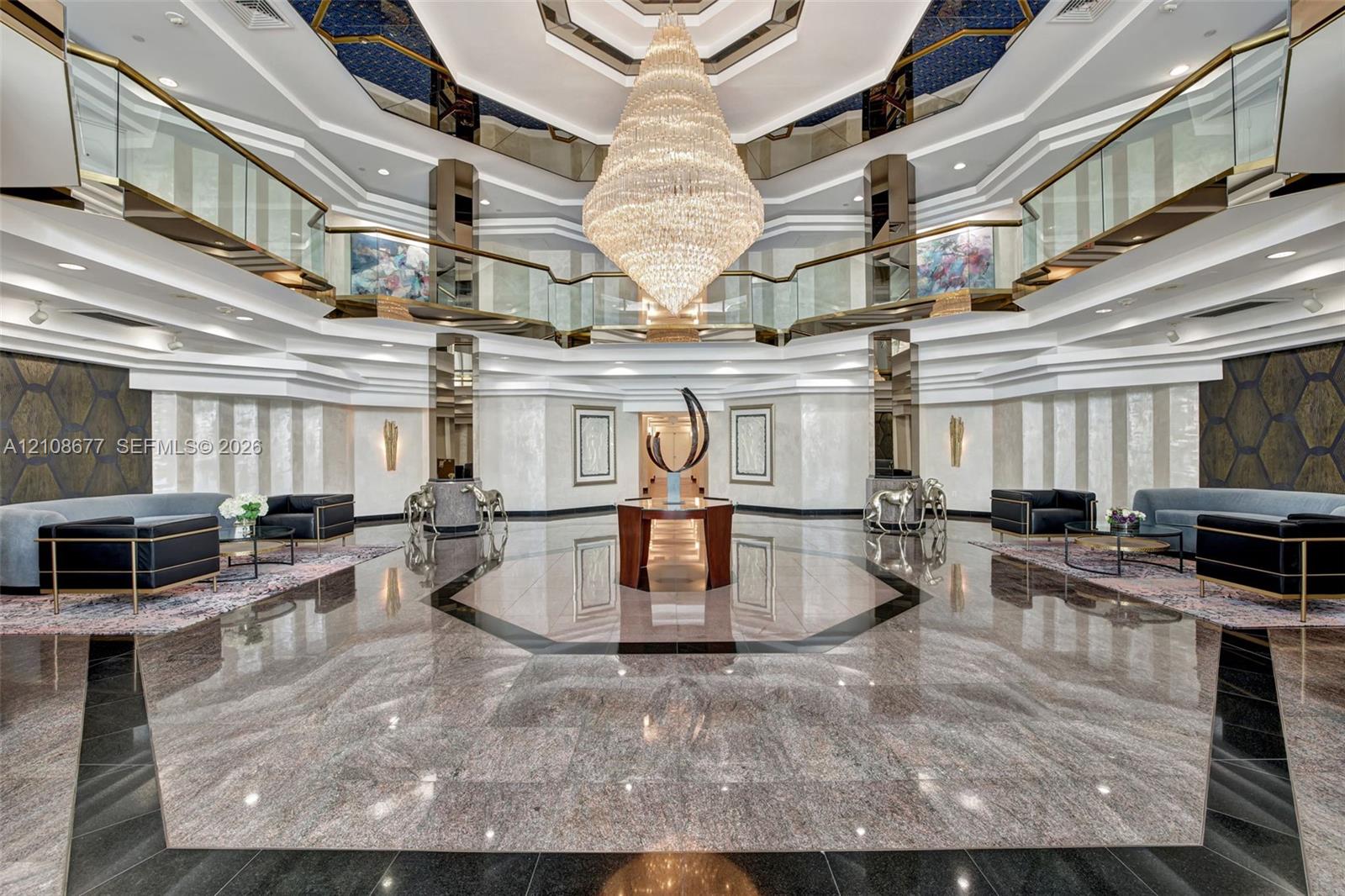 Grand lobby foyer featuring an expansive polished stone floor, a suspended multi-tiered crystal chandelier, a multi-faceted coffered ceiling with recessed lighting, and a wrap-around glass-paneled mezzanine and newer lobby furniture