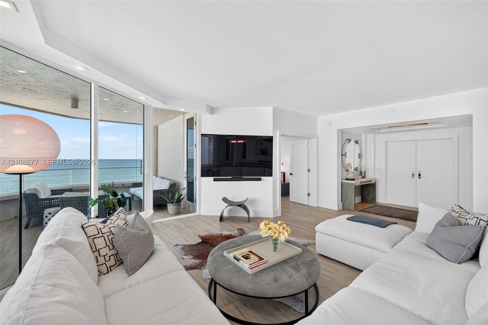 Living area featuring floor-to-ceiling glass panel walls, wood look plank porcelain flooring, integrated lighting, and a glass-railed balcony for more ocean views. TV wall was expanded to accommodate a larger TV. Light filtering remote operated shades with Bandalux