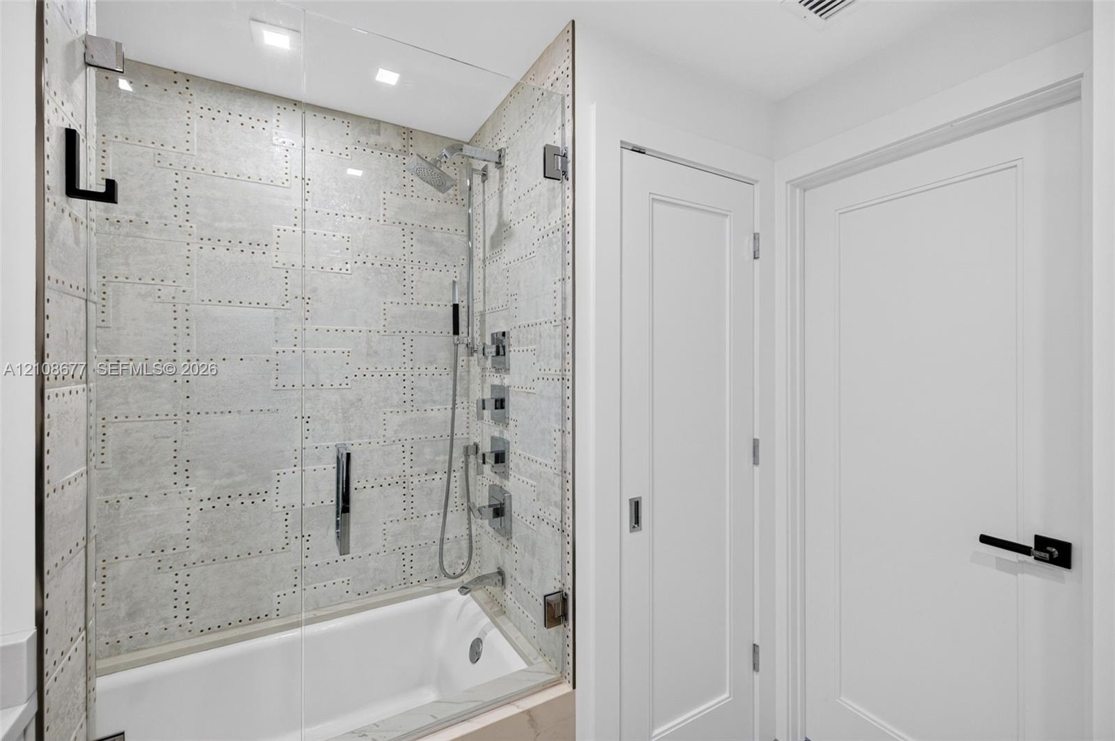 Shower enclosure featuring luxury patterned wall tile, frameless glass door, adjustable-height handheld showerhead, wall-mounted faucet controls, and recessed ceiling lighting plus linen closet