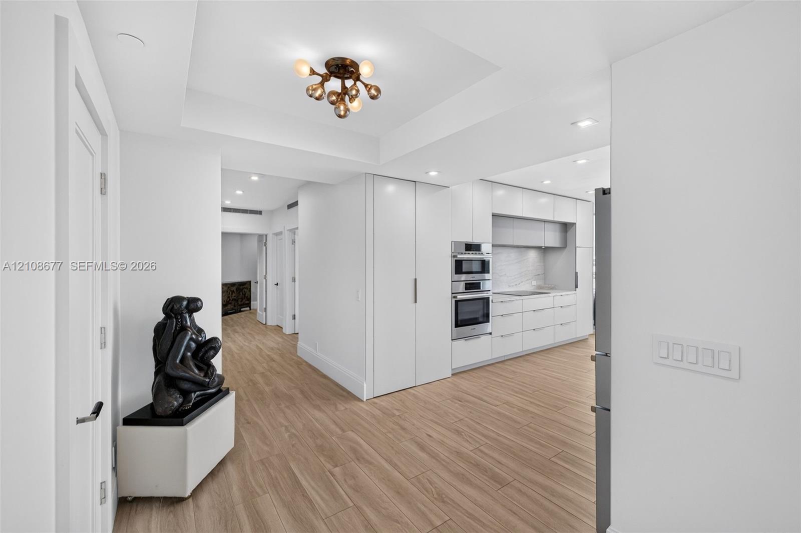 Widened hallway with primary bedroom straight down the hall, second bedroom next to it, plus gorgeous modern kitchen featuring white flat-panel cabinets, a gorgeous architectural multi-bulb flush-mount fixture, built-in Bosch stainless double ovens, and wood look plank porcelain flooring