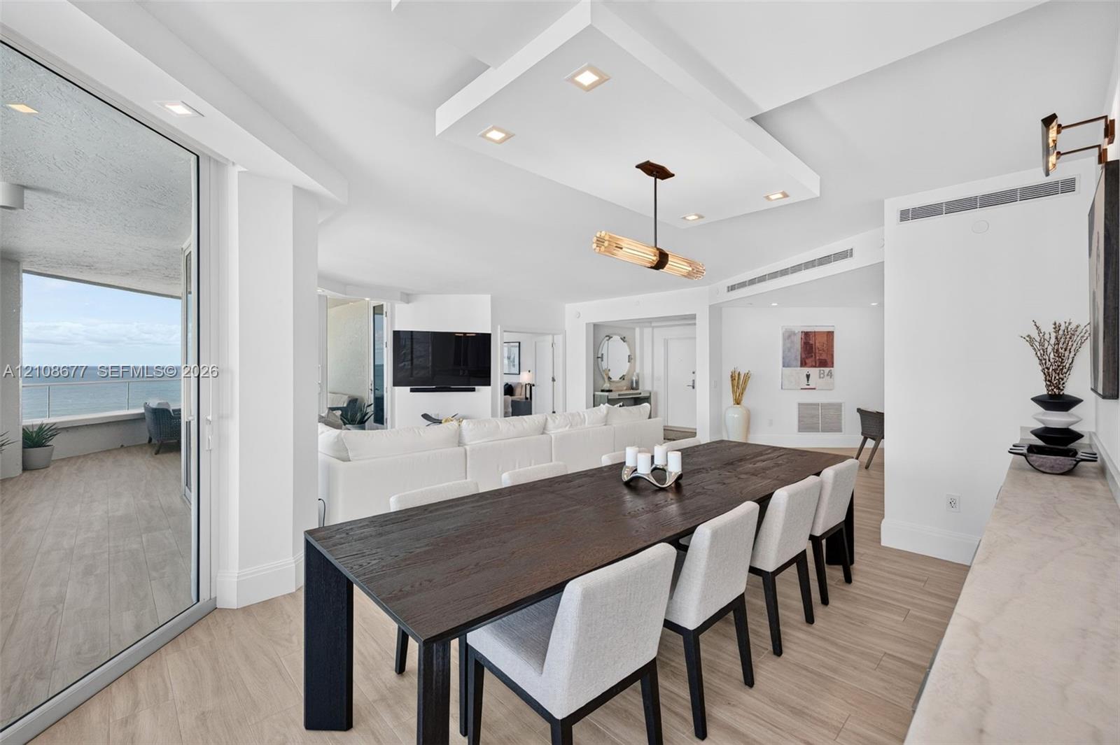 Open-concept interior featuring wood look plank porcelain flooring, recessed lighting, a custom architectural ceiling, a modern linear chandelier, and floor-to-ceiling glass doors opening to an exterior balcony from dining area w/ built in custom buffet for extra storage. Sliders lead again to the balcony for entertaining