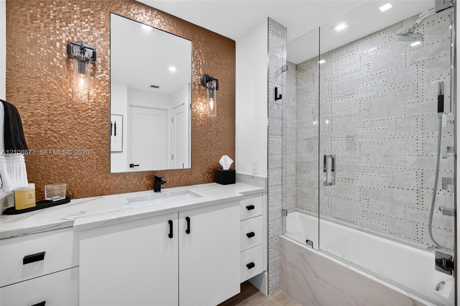 Bathroom featuring a hammered metallic tile accent wall, white floating vanity with stone counters, undermount sink, frameless glass shower enclosure, and custom decorative wall tile. Tub / shower combo w/ handheld shower head & rain head