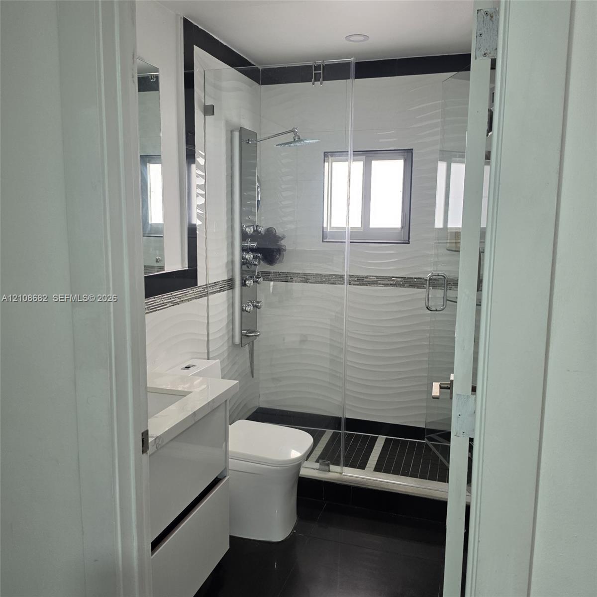 Bathroom featuring a wall-mounted shower panel system, glass shower enclosure, vanity with a stone-top counter, textured tile wall accents, and dark floor tiling