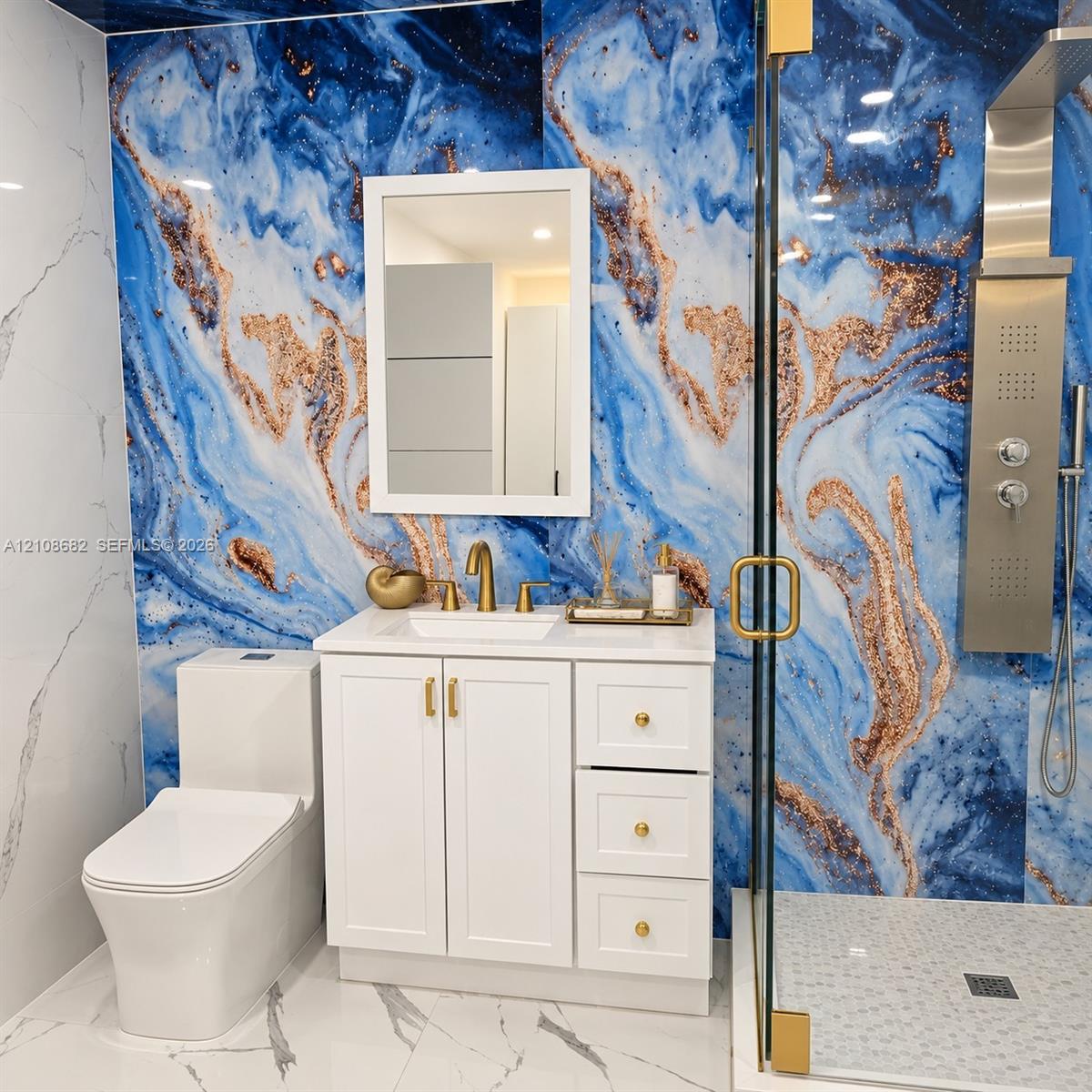 Bathroom featuring high-contrast blue and copper-toned wall paneling, white vanity with gold-finish hardware, rectangular under-mount sink, and a glass-enclosed shower with a metallic multi-function shower panel