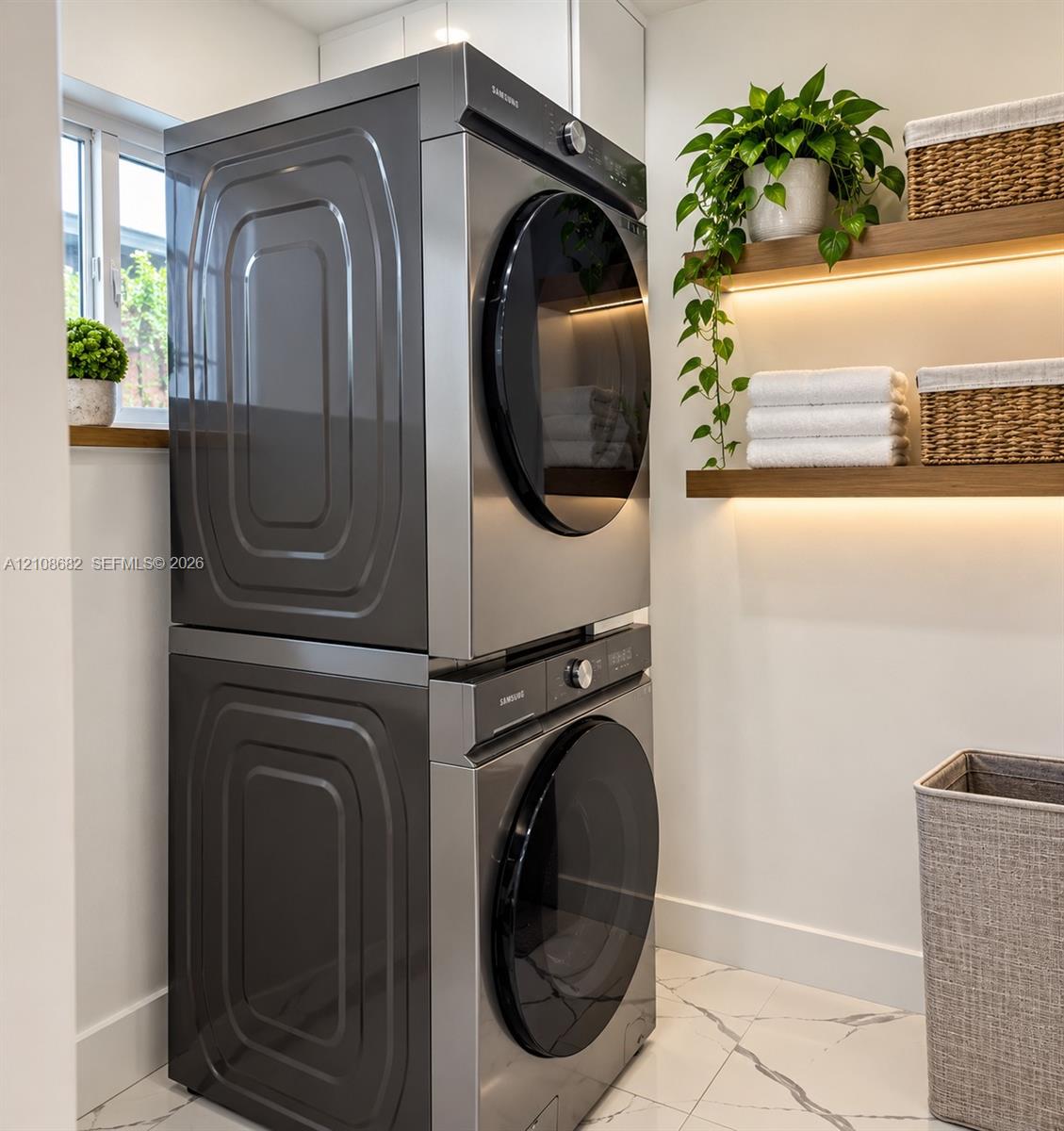 Stacked stainless steel laundry appliances, floating wooden wall shelves with integrated lighting, and white flooring with dark veining
