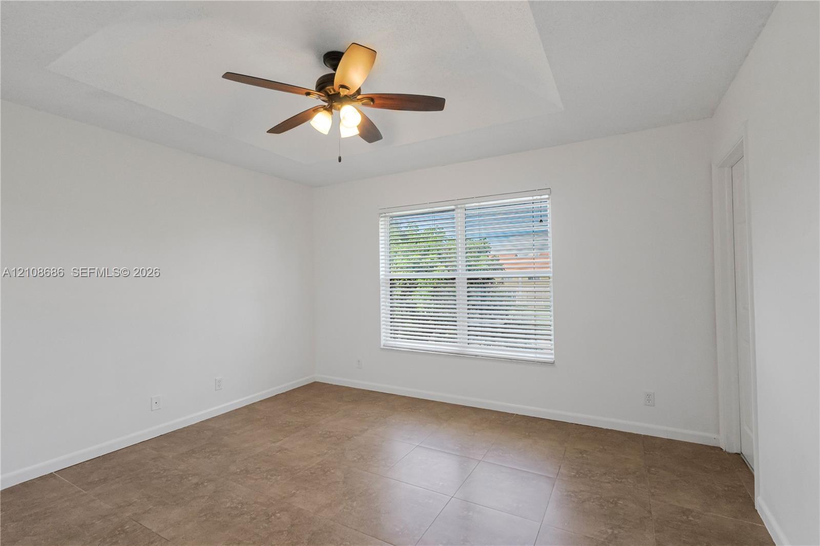 Room featuring a tray ceiling, wood-finish ceiling fan with integrated lighting, tile flooring, and a window with horizontal blinds Room featuring a tray ceiling, wood-finish ceiling fan with integrated lighting, tile flooring, and a window with horizontal blinds