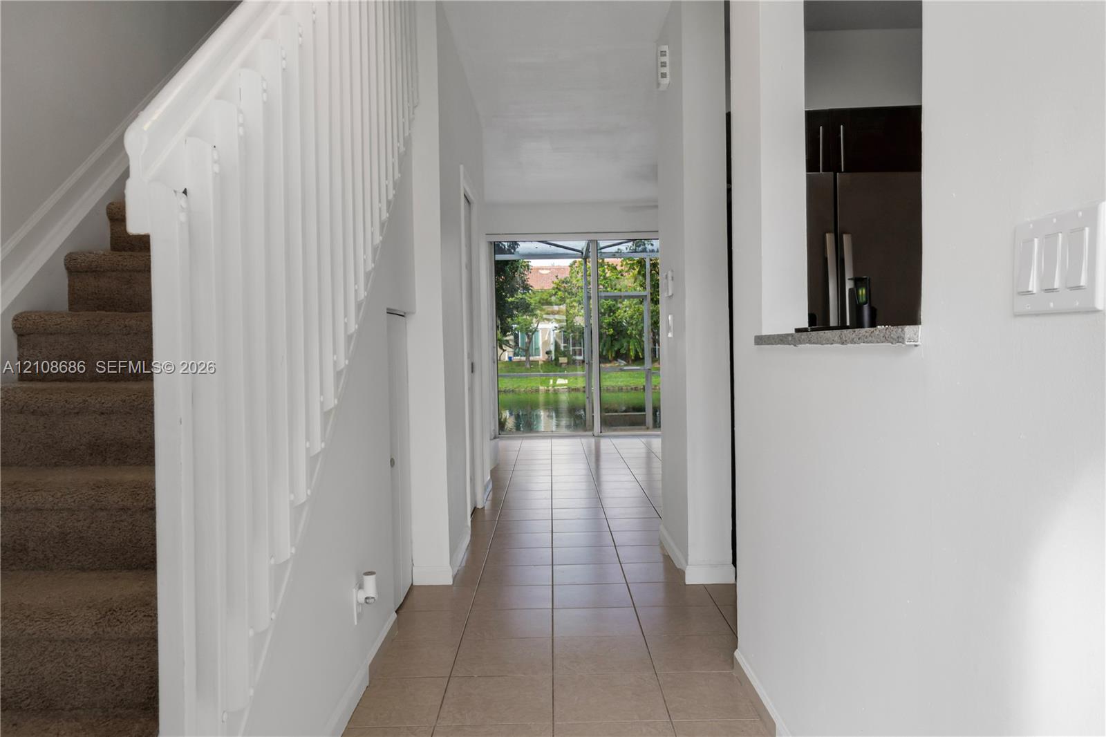 Interior hallway featuring ceramic tile flooring, a staircase with a white balustrade, and a pass-through opening to the kitchen area Interior hallway featuring ceramic tile flooring, a staircase with a white balustrade, and a pass-through opening to the kitchen area