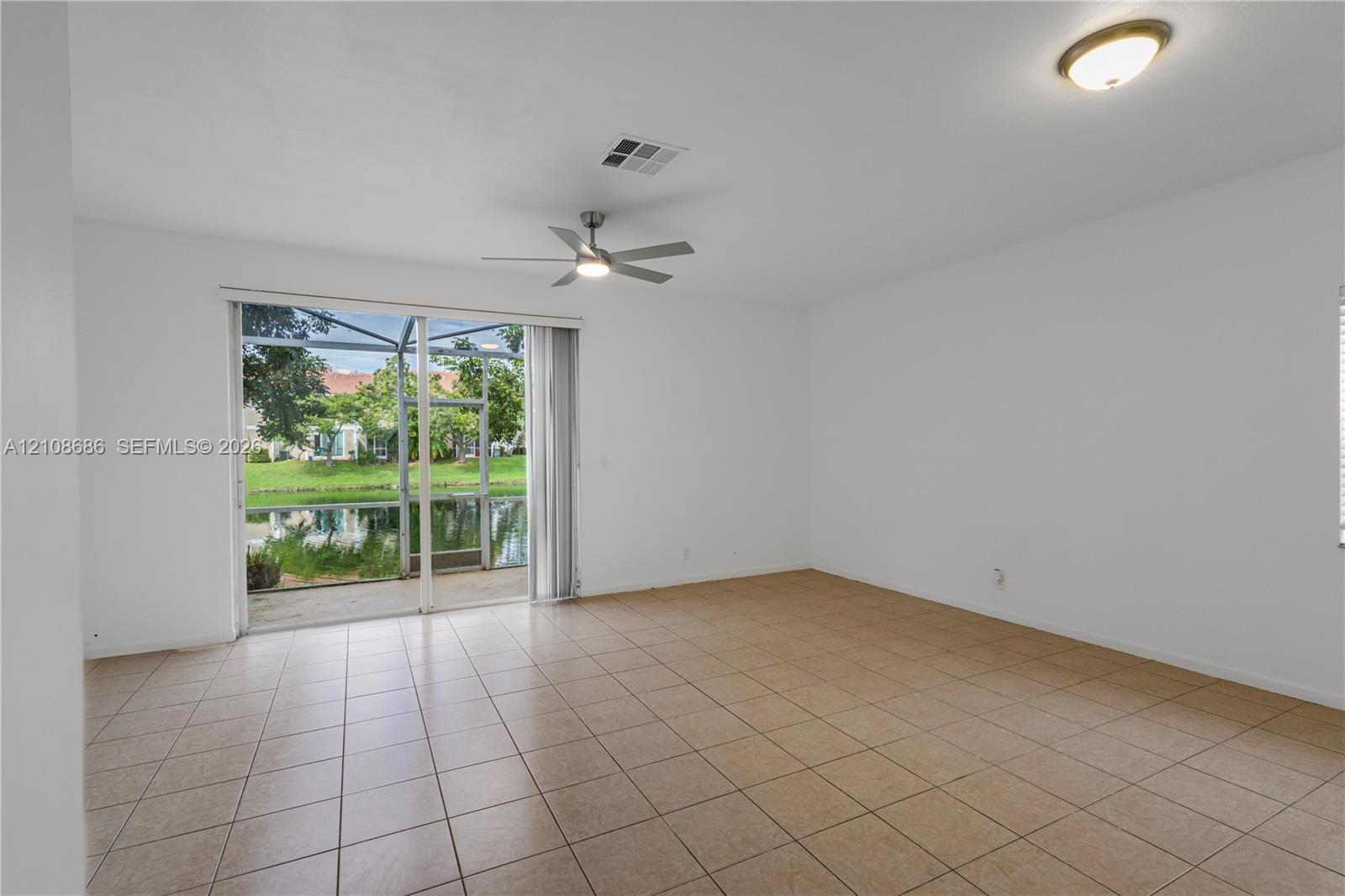 Open-concept interior featuring large-format tile flooring, a central ceiling fan, and sliding glass doors providing access to a screened-in patio overlooking a pond Open-concept interior featuring large-format tile flooring, a central ceiling fan, and sliding glass doors providing access to a screened-in patio overlooking a pond