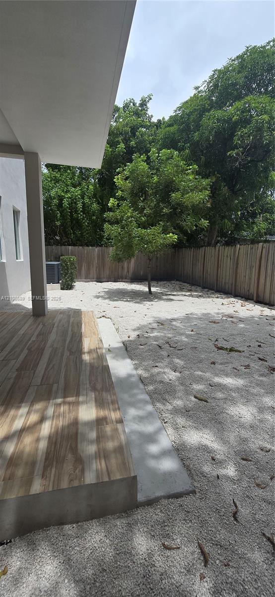 Covered patio with wood-finish tile flooring, fenced outdoor space with aggregate ground cover, and mature trees