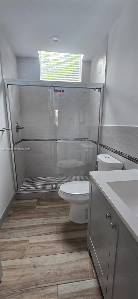 Walk-in shower with sliding glass door, tiled wall surround, skylight, wood-finish flooring, and vanity with solid surface countertop Walk-in shower with sliding glass door, tiled wall surround, skylight, wood-finish flooring, and vanity with solid surface countertop